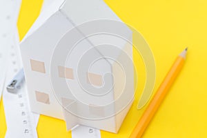 White paper house, yellow pencil and white measuring tapes with centimetre and inches on vivid yellow background, architecture,