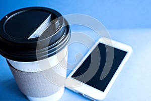 White paper kraft disposable cup for coffee with black plastic lid and white mobile phone on blue background