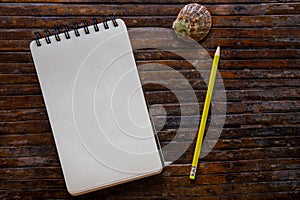 White paper notebook with pencil and shell on rustic wooden table top view photo. Vertical sketchpad with blank page