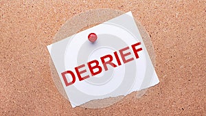 White paper with the text DEBRIEF is attached to the wooden background with a red button