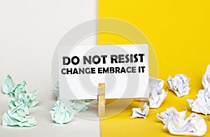 White paper with text Do Not Resist Change Embrace It on a clothespin on yellow and white backgrounds with paper wads of different