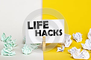 White paper with text Life Hacks on a clothespin on yellow and white backgrounds with paper wads of different colors