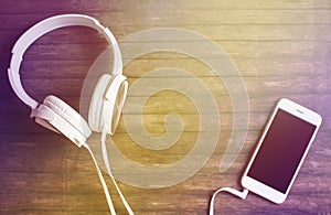 White phone and headphones on wooden table. Warm yellow light toned photo.