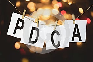 White pieces of paper with the text PDCA Plan Do Check Act hang on a rope against a multicolored bokeh background