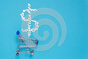 White pills in dollar sign shape over shopping cart on blue background. Pharmaceutical concept of medical expenses