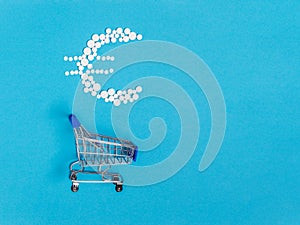 White pills in euro sign shape over shopping cart on blue background. Pharmaceutical concept of medical expenses