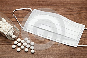 White pills spilling out of a transparent medicine bottle and mask on wood.