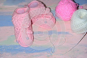 White-pink knitted baby shoes booties on a pink-blue wooden table with balls of thread and a heart of threads. Newborn