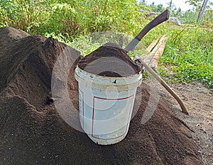 white plastic barrel containing ground manure. around it is a pile of fertilizer and two hoes