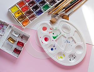 Palette with a set of watercolor paints and brushes on a pink background, the artist`s tools