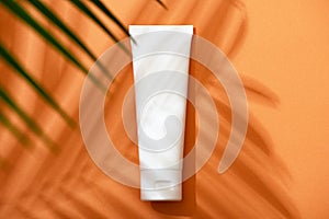 White plastic tube with face, hand and body cream on an orange background with palm leaves and shadow. Sun protection lotion,