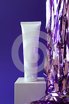 White plastic tube mockup with moisturizer cream, shampoo or facial cleanser and holiday tinsel on cube podium on violet