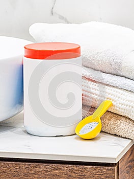White plastic washing powder container with red cap staying on bathroom table near fresh towels and yellow spoon