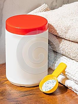 White plastic washing powder container with red cap staying on bathroom table near fresh towels and yellow spoon