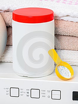 White plastic washing powder container with red cap staying on washing machine near fresh towels and yellow spoon