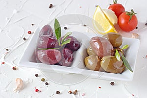 White plate with black and green olives with olive leaves on a white abstract background with lemon, cherry tomato