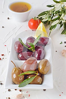 White plate with black and green olives with olive leaves on a white abstract background with lemon, cherry tomato