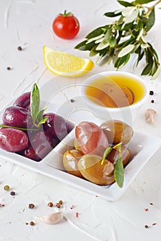 White plate with black and green olives with olive leaves on a white abstract background with lemon, cherry tomato