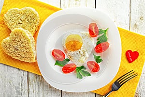 A white plate with cherry tomatoes, parsley, a heart-shaped fried egg and two slices of bread on a yellow napkin