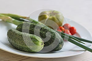 White Plate with fresh vegetables on wooden table. Two cucumbers, red cherry tomatoes, green cracked tomato with vertical splits,