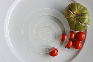 White Plate with fresh vegetables on wooden table. Two cucumbers, red cherry tomatoes, green cracked tomato with vertical splits,