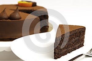 White plate with a piece of delicious chocolate cake and the cake in background