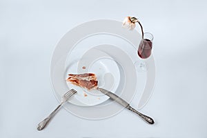 White plate with a piece of partially eaten pie, fork, knife and glass of champagne with faded rose on a white