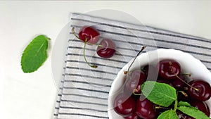 A white plate with a ripe burgundy cherry and a mint petal stands on a striped kitchen towel on a white background and