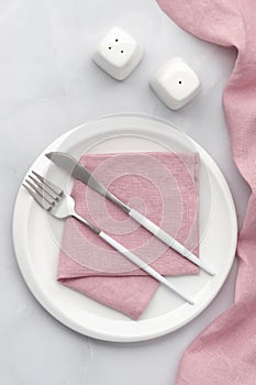 White plate with silver cutlery. Knife, fork and pink napkin on grey background. Table setting, flat lay, top view. Serving space