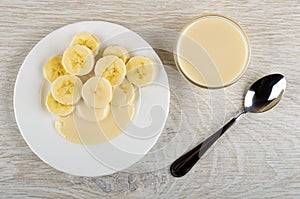 White plate with slices of banana and condensed milk, bowl with milk, spoon on wooden table. Top view