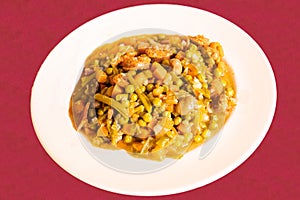 A white plate of vegetable stew with legumes such as peas, carrots, abas and chorizo on a red tablecloth