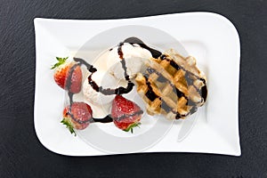 White plate with waffle, ice and strawberries decorated with chocolate stripes on a dark slate