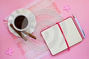 White porcelain cup with coffee, two coffee rolls on saucer with wavy edge and paper notebook with ball pen on a pink table napkin