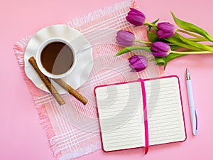 Porcelain cup with coffee and two coffee rols on saucer with wavy edge, paper notebook with ball pen and bouquet of pink tulips on