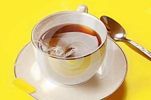 White porcelain cup with hot freshly brewed tea, bag still in, silver spoon next on yellow board, lit by morning sun