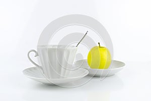 White porcelain cup with spoon and saucer, yellow apple on saucer, on white surface.