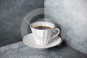 White porcelain hot coffee cup with spoon and saucer on gray surface in the corner