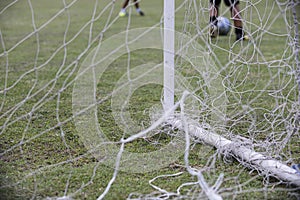 A white post of football goal with a white broken and messy net, while boy`s game playing on.