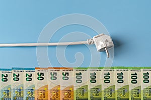 White power plug above row of euro money banknotes against blue background. Concept of rising energy bills and electricity price