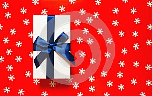 White present box with dark blue ribbon and bow on red background with decorative snowflakes. Christmas gift concept. Festive and