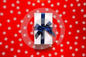 White present box with dark blue ribbon and bow on red background with decorative snowflakes. Christmas gift concept. Festive and