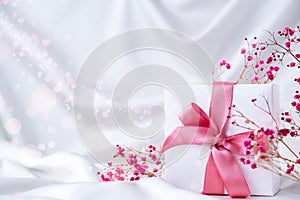 White present box with pink ribbon and small pink flowers on white silk fabric background. Greeting card for holidays