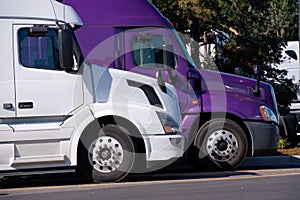 White and purple big rigs semi trucks stand on truck stop side b