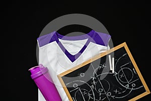 White and Purple Sports Uniform and Water Bottle Next to a Chalkboard on Black Background