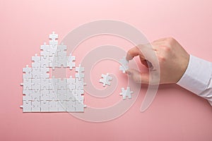 White puzzle. House shape puzzle. The concept of rent, mortgage. Hand holding piece of white puzzle. Pink background