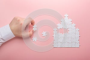 White puzzle. House shape puzzle. The concept of rent, mortgage. Hand holding piece of white puzzle. Pink background