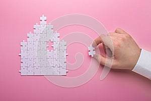 White puzzle. House shape puzzle. The concept of rent, mortgage. Hand holding piece of white puzzle. Pink background