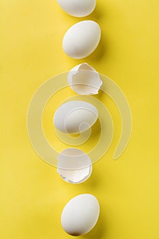 White raw chicken eggs lying in vertical row with broken egg on yellow background.