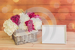 White and red peony flowers in wicker basket and white frame on wooden table.