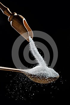 White refined sugar crystals falling down into the wooden spoon at black background